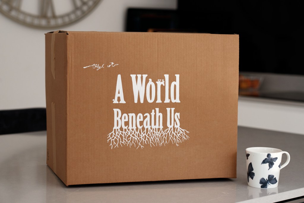 A brown cardboard box sits on a desk with a coffee mug next to it. On the box is the title of the show: "A World Beneath Us" in bold white font. The words "Beneath Us" are stylised to have what looks like roots growing below them.