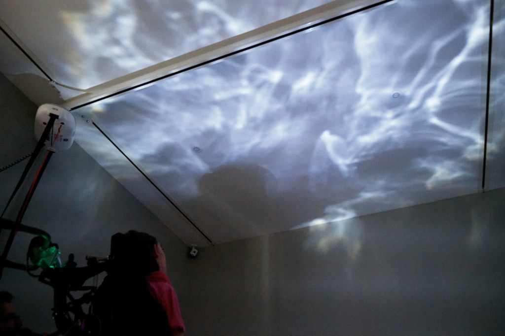 A child using a wheelchair looks up towards the ceiling, where lights are projected. The lights are white, and look like they are moving, almost like water