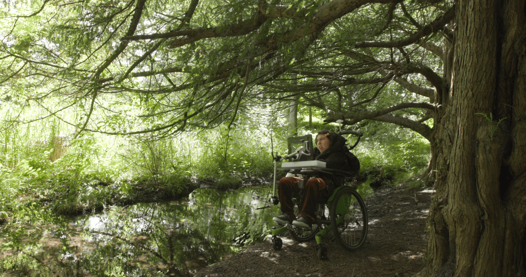 A still from the film accompanying "A World Beneath Us".
A wheelchair user sits by a body of water in a forest