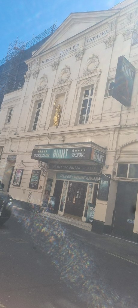 The facade of the Harold Pinter theatre - a white building with a gold statue between the theatre windows. The signage for the play: Giant features a blue- green background and two five star reviews bookend the title. All the text is in bold, white lettering, with the "A" in "Giant" artfully torn.